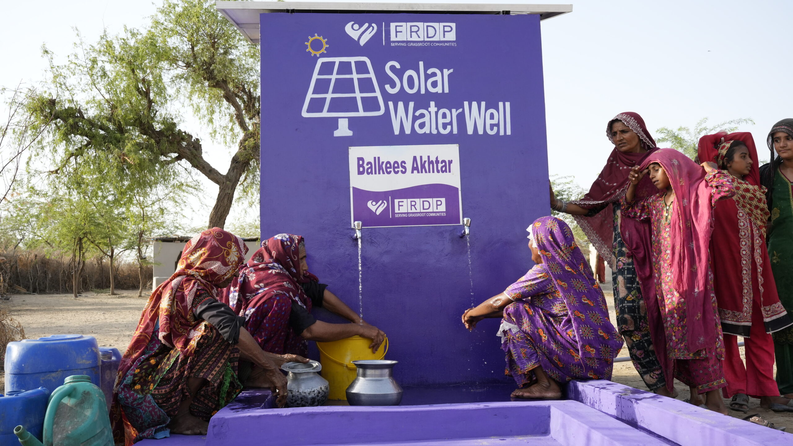 climate-change Women and children in colorful traditional clothing collect water from a purple solar-powered water well labeled “Solar Water Well Balkees Akhtar FRDP” in a rural outdoor setting.