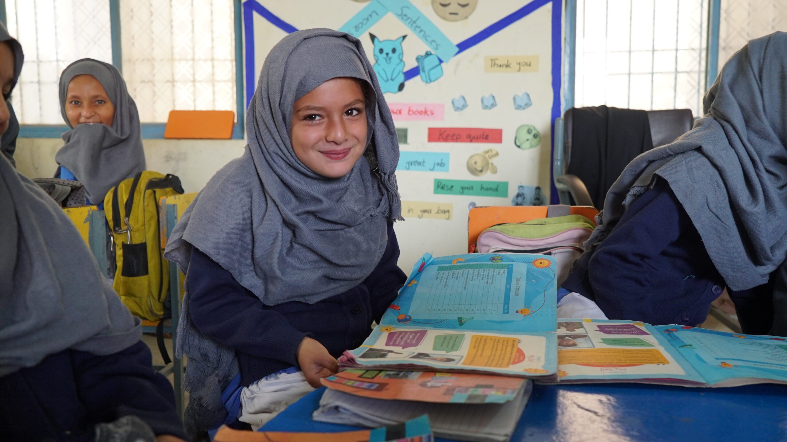 education-and-awareness A young girl wearing a gray headscarf and navy uniform smiles while sitting at a desk with open books in a colorful classroom, alongside other students.