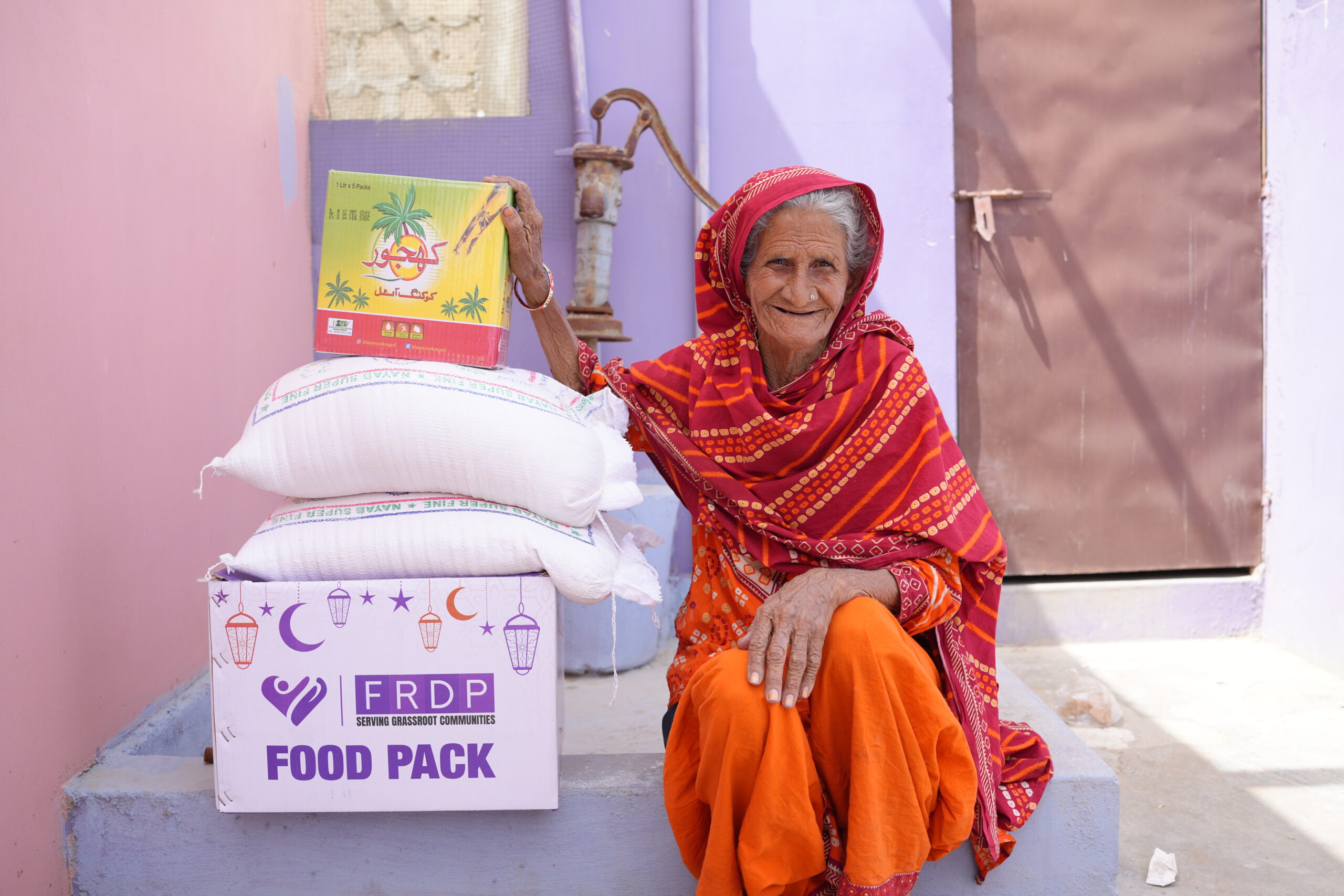 emergencies-disaster-risk An elderly woman in colorful traditional attire sits smiling next to white bags and a box labeled FRDP Food Pack, holding a food item, with pastel-colored walls in the background.