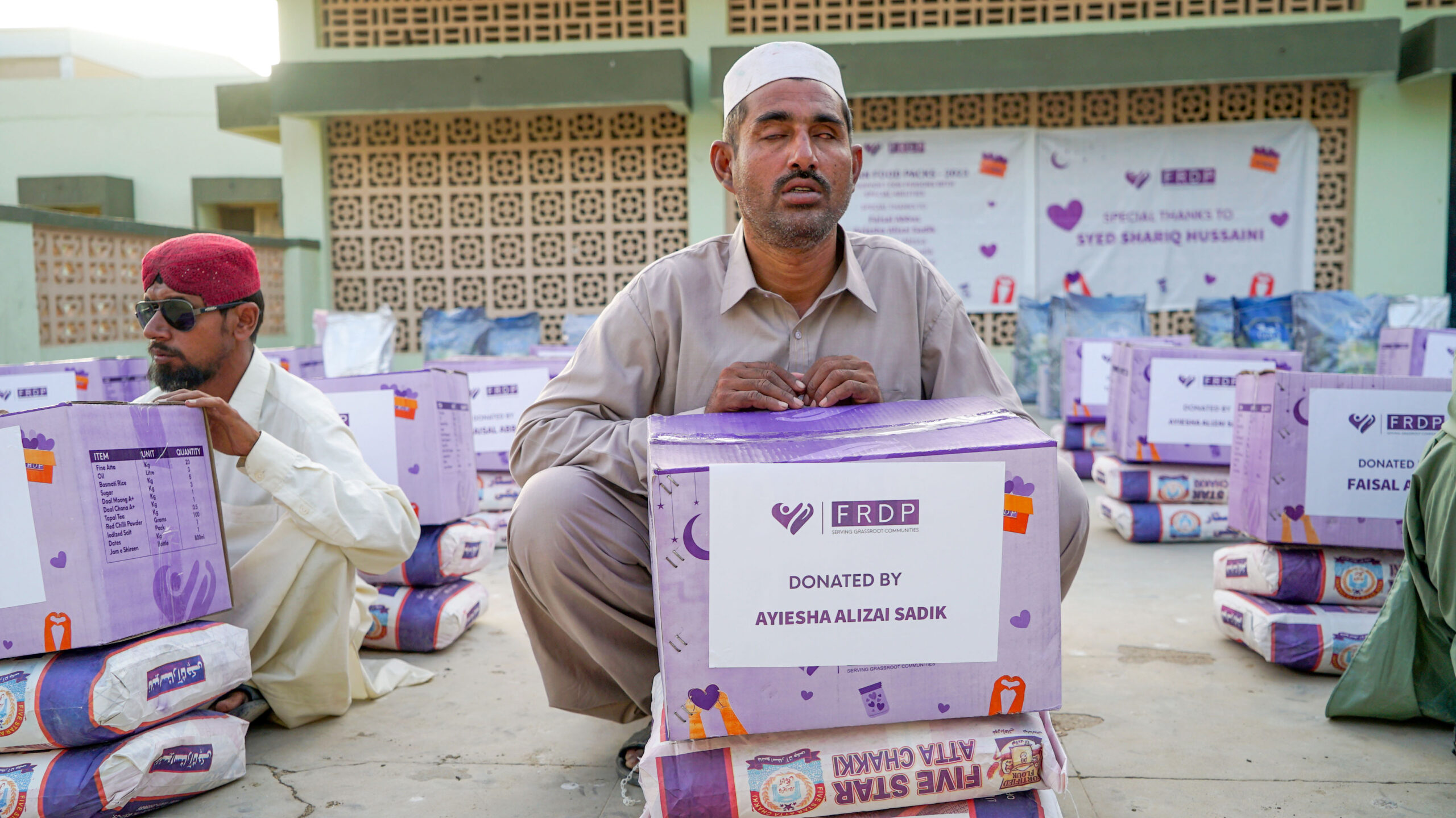 food-security-nutrition A man in traditional clothing sits outdoors with a large donation box labeled FRDP in front of him. Several similar boxes and people are visible around him, with a banner in the background.
