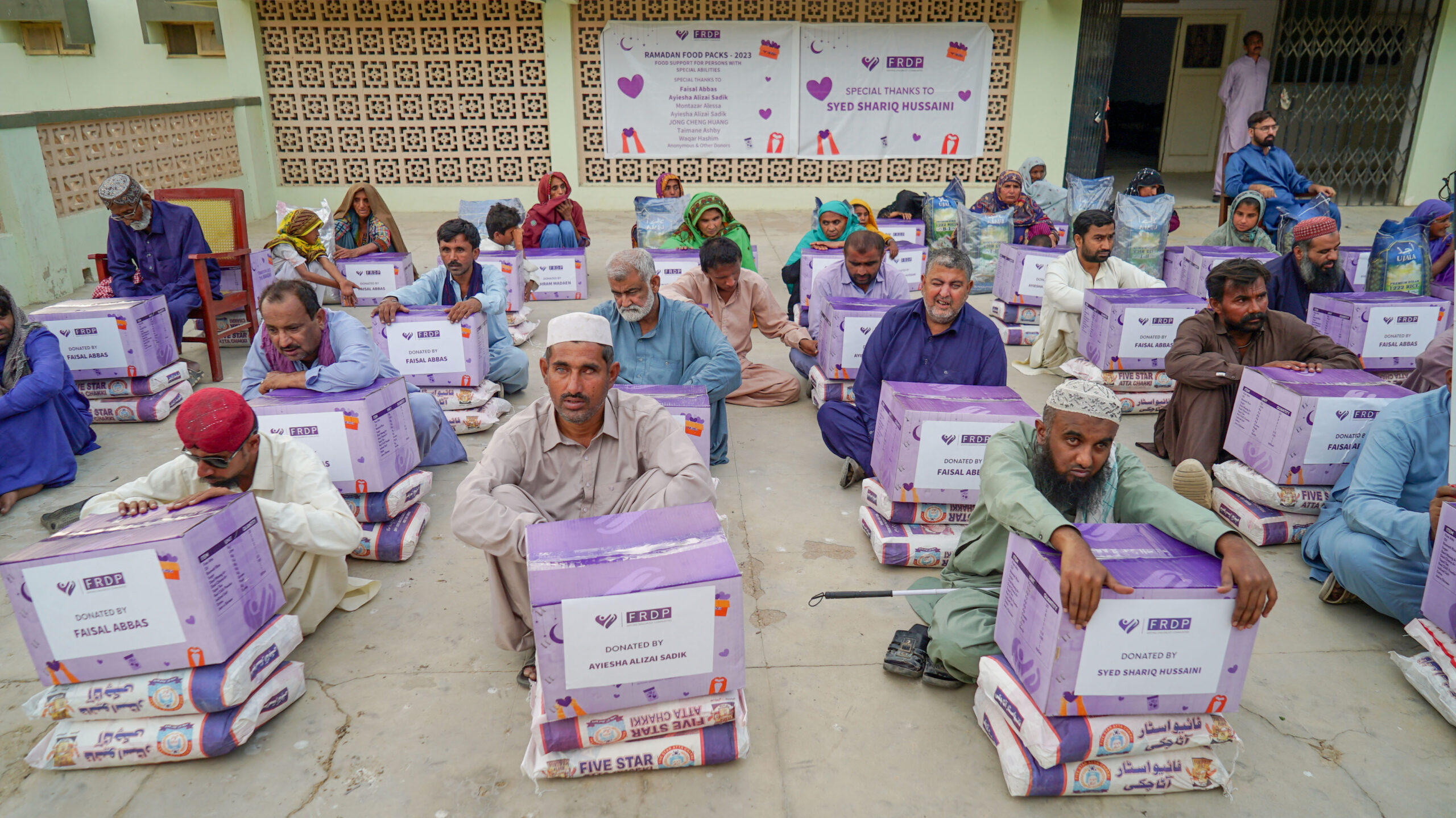 AppealforFood A group of men in traditional clothing sit on the ground, each with a purple food aid box and a sack of flour, in front of a building with banners displaying logos and messages of gratitude.